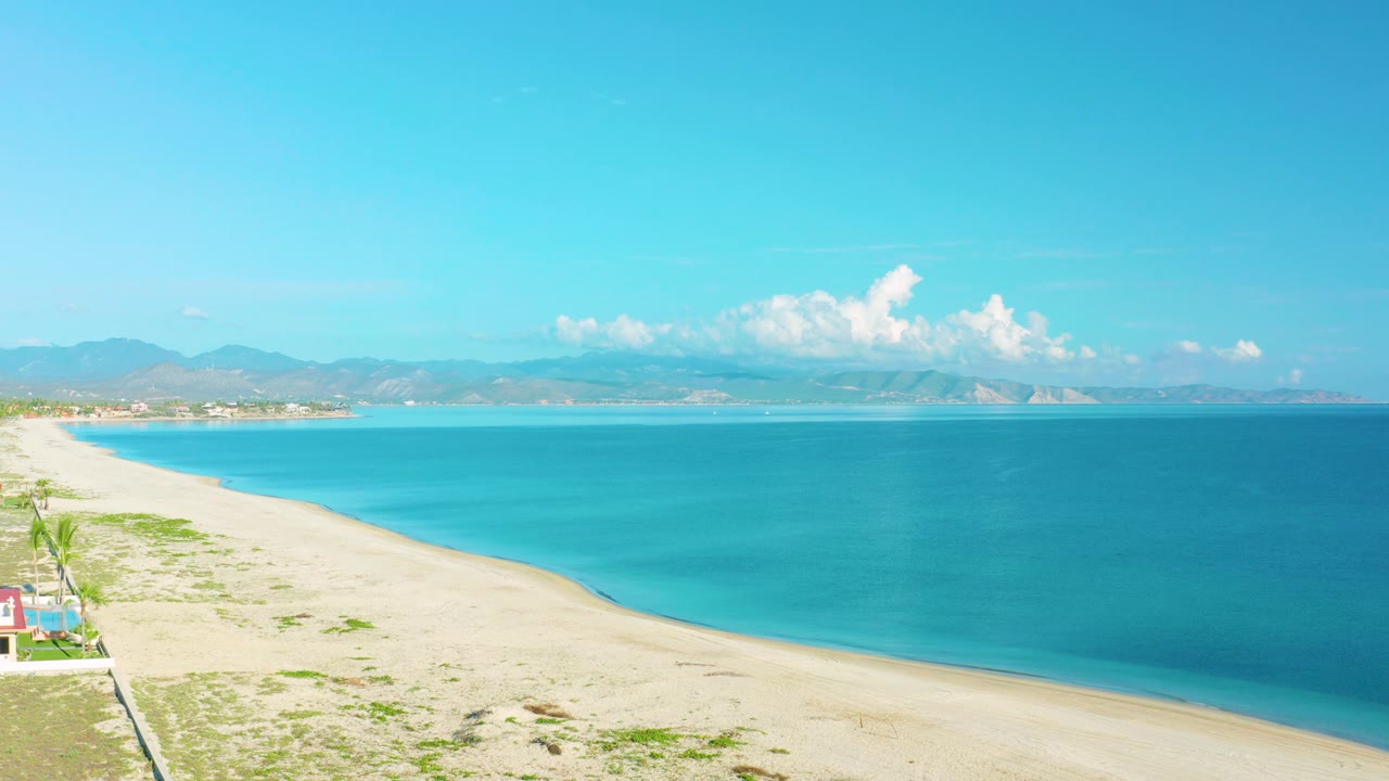 Aerial beach scene with sea, sand, vegetation, and mountains under blue sky.