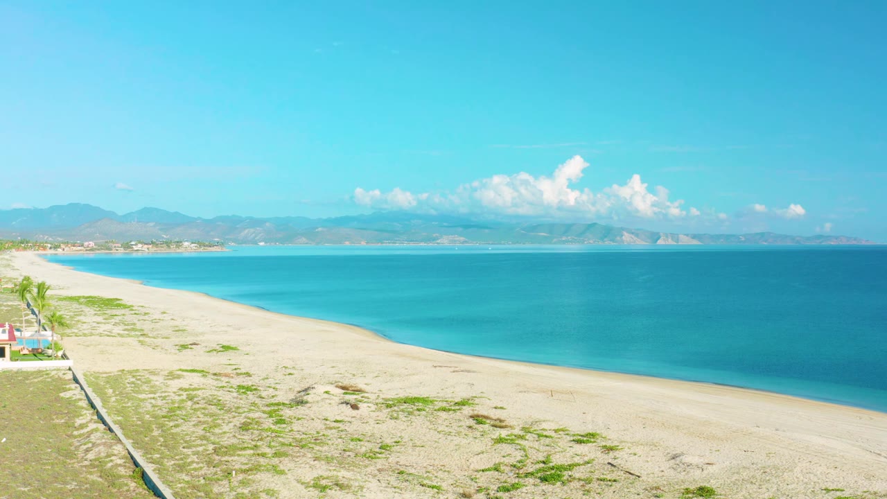 La Ribera coastline at 0:01, showing a long pale beach, blue-green water, faint surf, mountains in the distance, and no visible people.