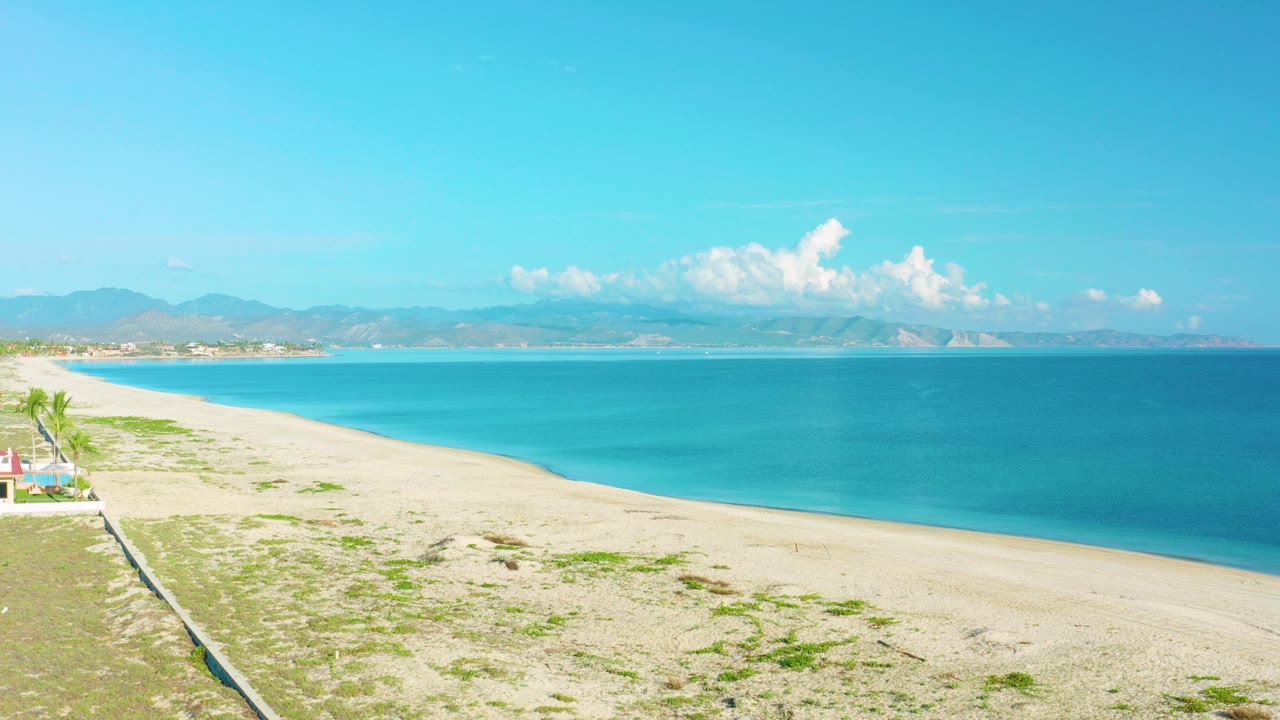Frame from the La Ribera clip with the beach stretching diagonally below, turquoise sea filling the middle of the view, distant mountains, and no visible people.