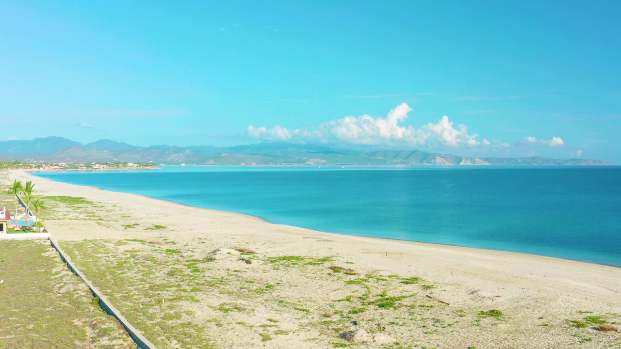 La Ribera beach view at 0:03 with a long strip of sand, calm blue ocean, mountain ridges across the horizon, and no visible people.