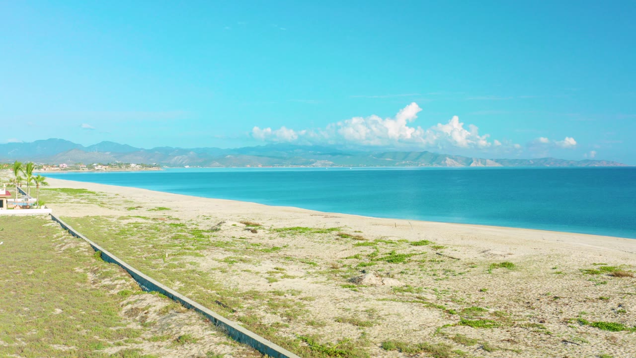 Wide elevated view of La Ribera beach with pale sand, blue sea, distant mountains, a low stone wall at left, and no visible people.