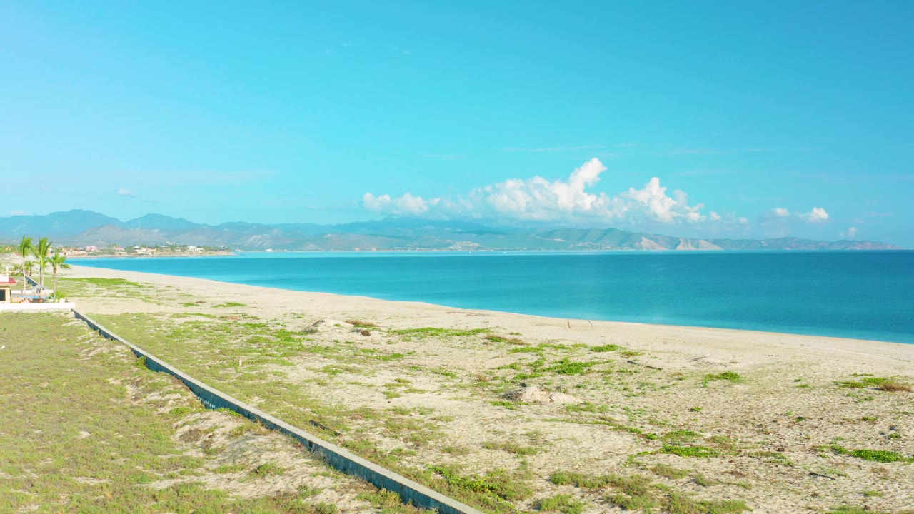 Wide La Ribera beach frame with the low wall leading from the foreground, open sand, turquoise sea, distant hills, and no visible people.