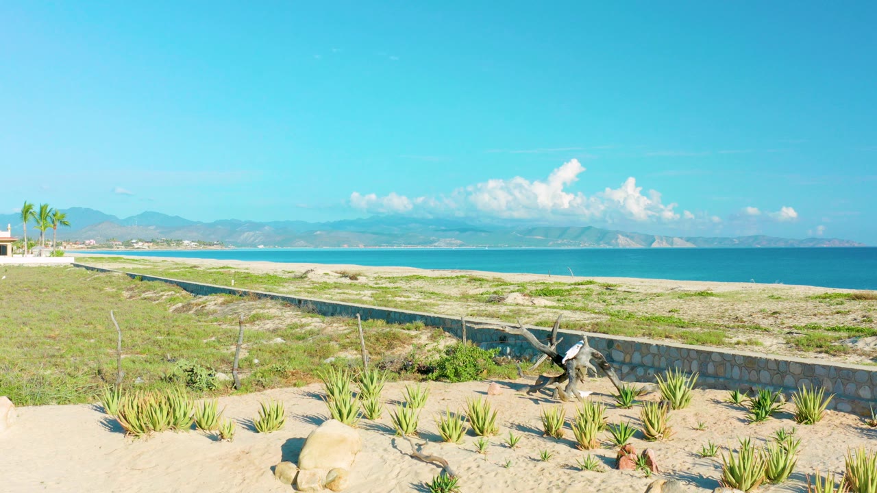 Wide La Ribera beach view with sand, rocks, desert plants, a low wall, turquoise water, distant mountains, white clouds, and no visible people at 0:00.