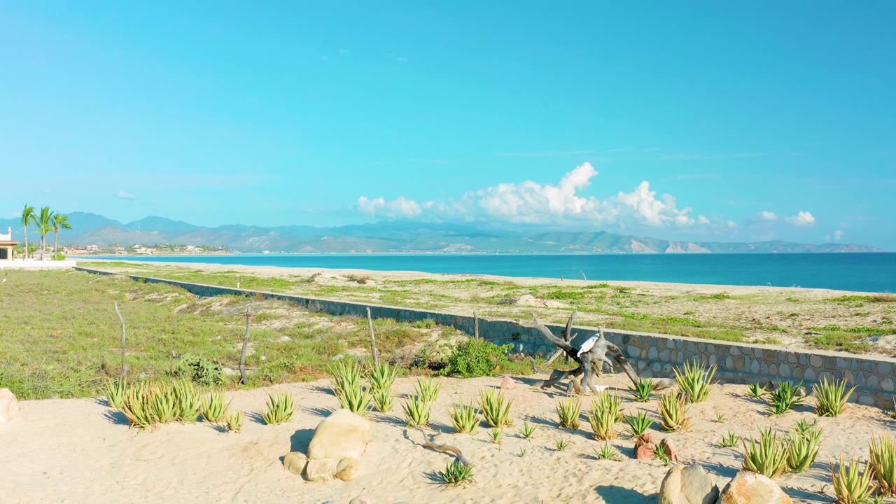 Wide La Ribera beach frame showing pale sand, scattered plants, a concrete wall, calm blue sea, mountain ridge, cloud bank, and no visible people at 0:02.
