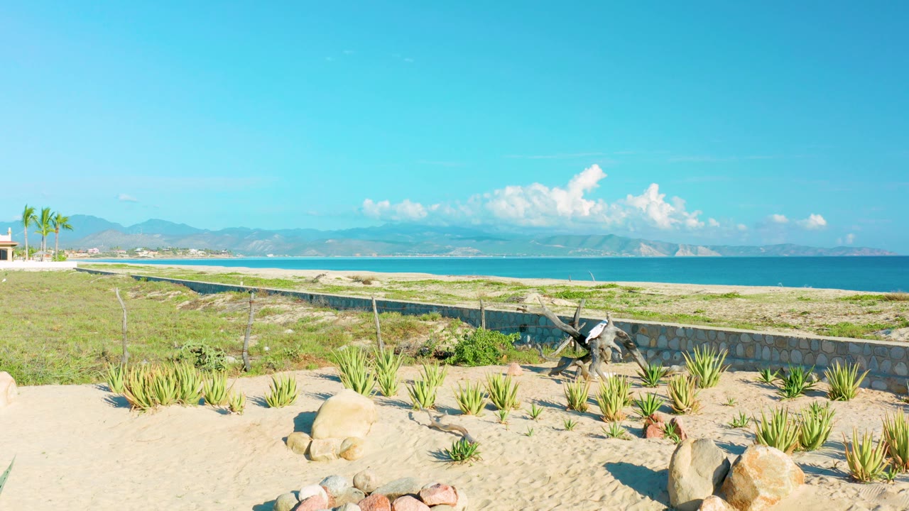 La Ribera beach view with more sandy foreground visible, low green plants, rocks, a wall, blue water, distant mountains, white cloud shapes, and no visible people at 0:03.