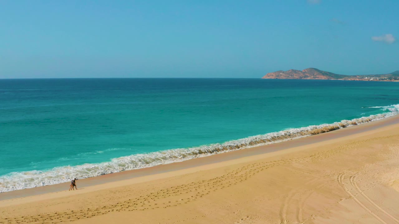 Two small figures walk together near the lower left surf edge at Dreams Beach, while white water rolls onto the sand and a low coastline sits across the horizon.