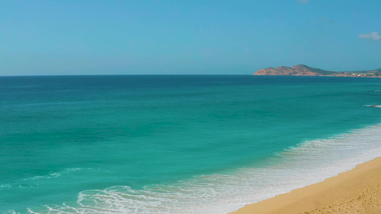 Blue-green sea with foamy shore break and low land across the horizon.