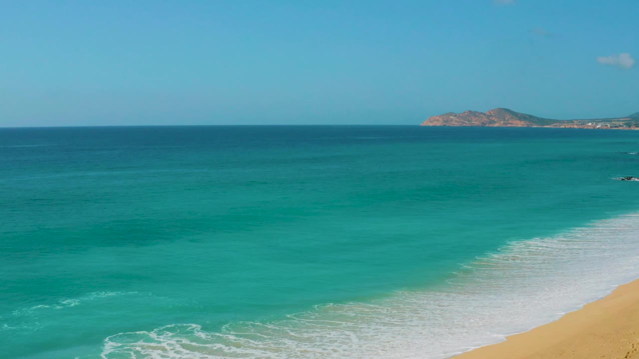 Dreams Beach ocean view with more open turquoise water across the center, a diagonal band of white surf entering from the right, distant rocky shoreline, and no visible people.