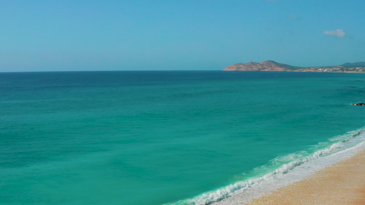 Wide view of turquoise water at Dreams Beach, Baja California Sur, with white surf reaching the sand, a distant rocky headland, pale sky, and no visible people.