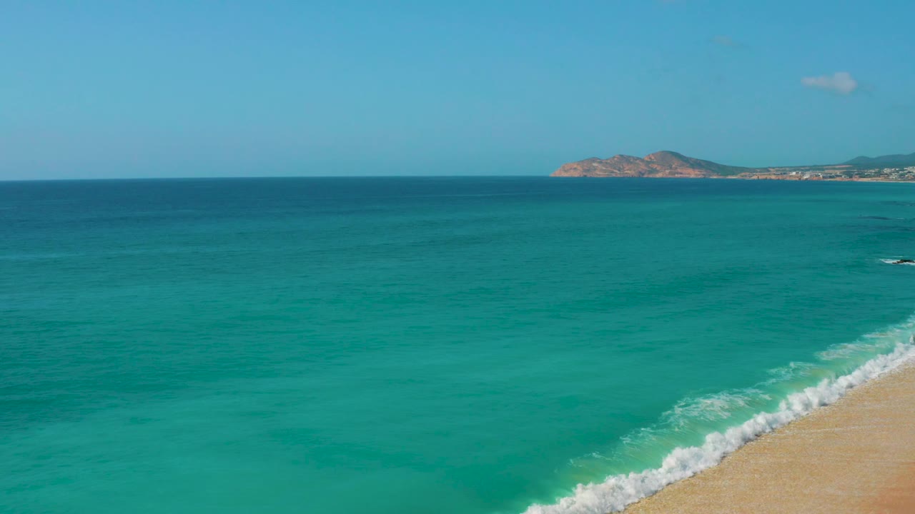 Clear turquoise ocean at Dreams Beach, Baja California Sur, with the surf angled across the lower right corner, a rocky headland in the distance, and no visible people.