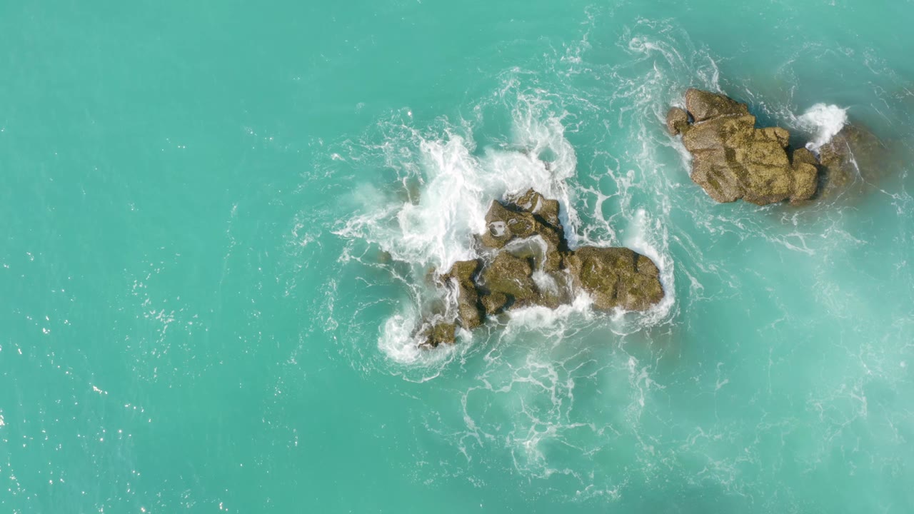 Aerial frame from Dreams Beach in Baja California Sur, Mexico, with white wave wash curling around the central rocks and a separate rock cluster near the upper right, with no visible people.
