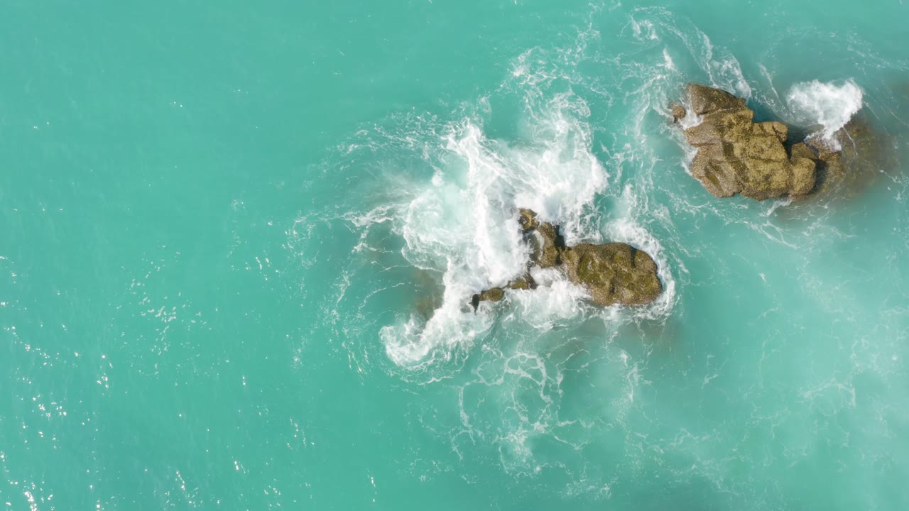 Top-down Dreams Beach view in Baja California Sur, Mexico, where a larger white splash spreads across the central rocks while the upper-right rock remains partly ringed by foam, with no visible people.