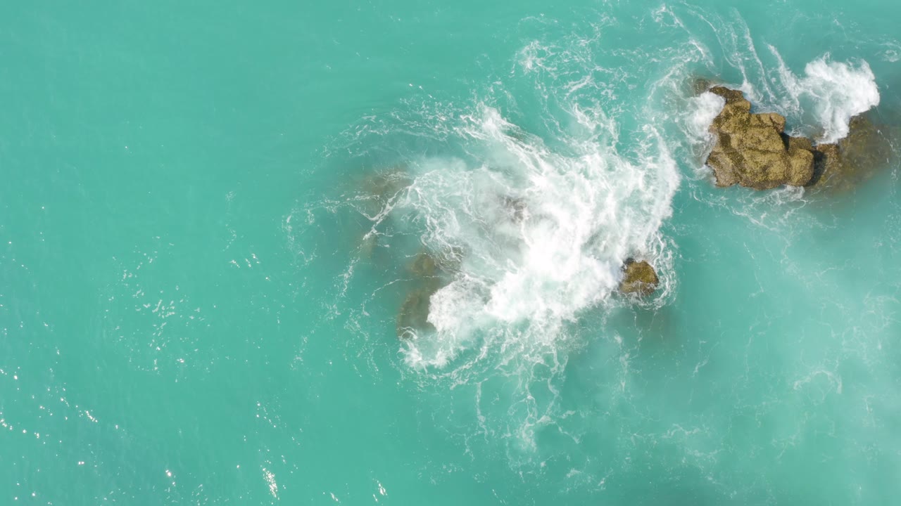 Overhead frame at Dreams Beach in Baja California Sur, Mexico, with dense white surf covering much of the lower rock group and a larger rock cluster sitting in clearer water above, with no visible people.