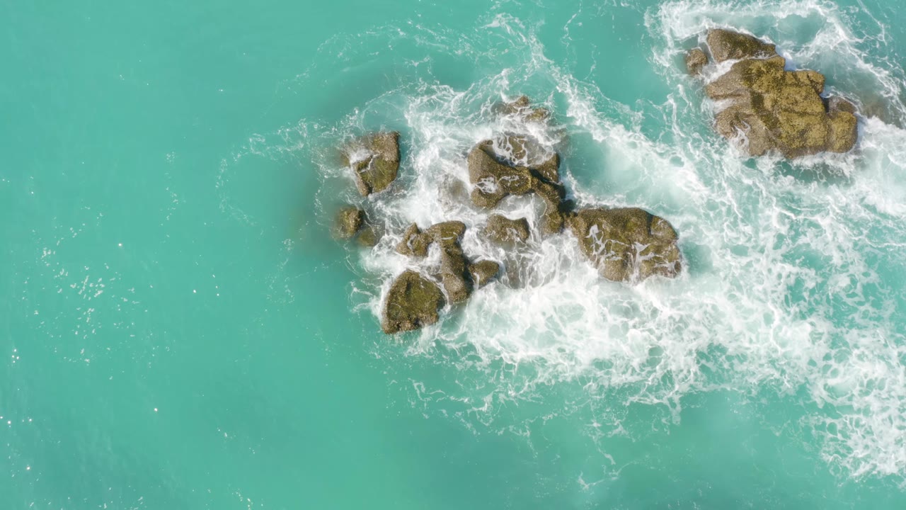 Aerial frame from Dreams Beach in Baja California Sur, Mexico, with white foam gathering along the right side of the central rock group and a separate rock near the upper right, no visible people.