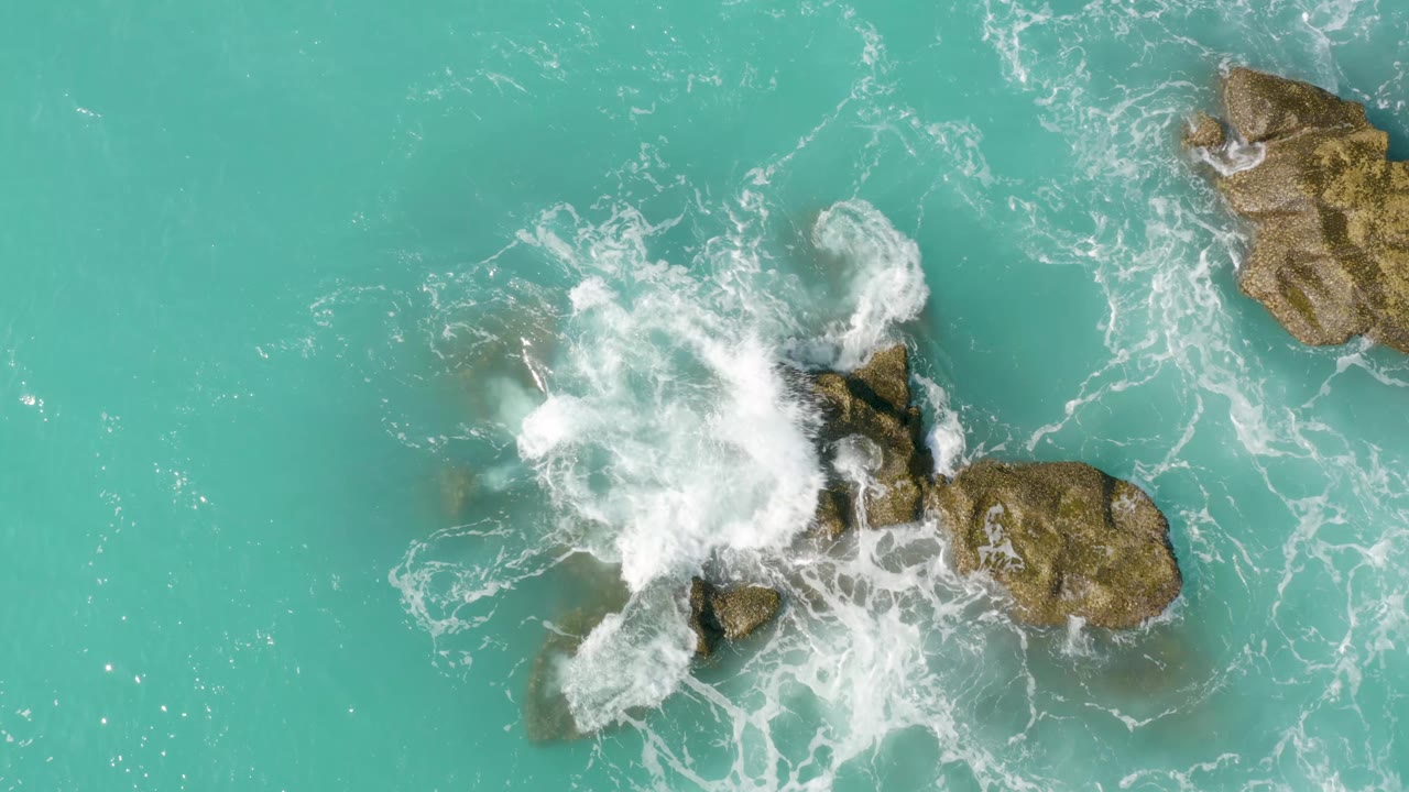 Aerial frame of whitewater spreading wider around several brown rocks in turquoise sea at Dreams Beach, with no visible people or structures.
