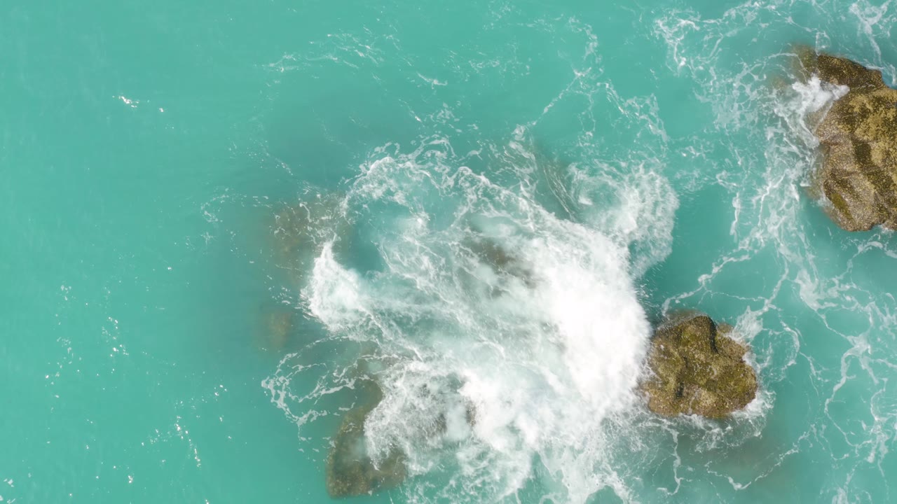 Top-down view of a larger burst of white foam covering part of the rock group in the turquoise water at Dreams Beach, with no visible people.