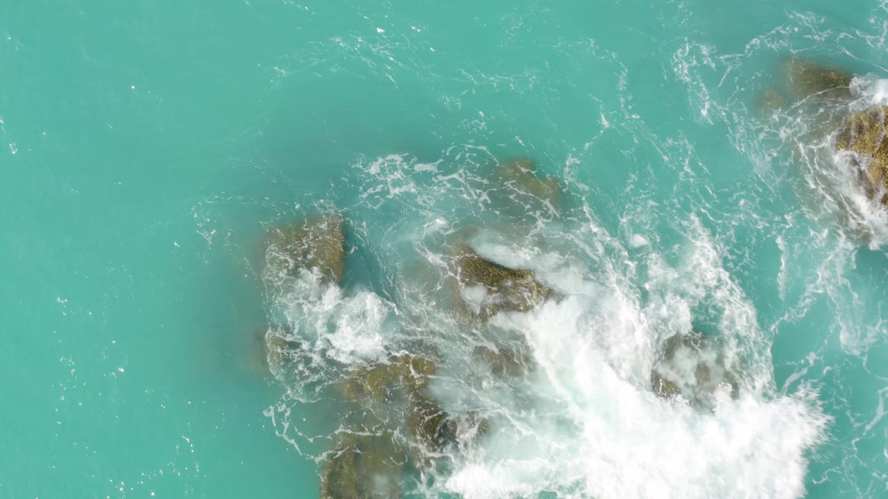 Overhead frame with the rocky surf zone shifted toward the lower edge, showing turquoise water above and whitewater churning across the rocks at Dreams Beach, no visible people.