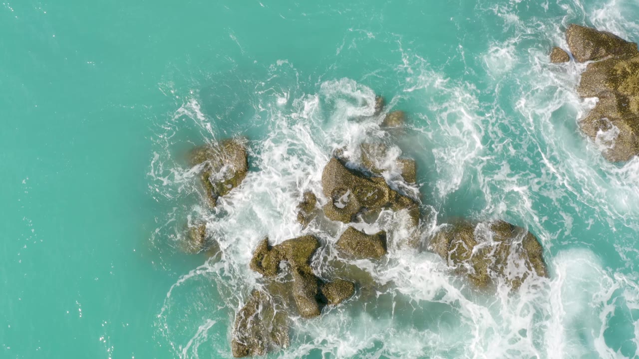 A larger patch of white water churns around the rock cluster in the turquoise sea at Dreams Beach, Baja California Sur, Mexico, with no boats or visible people.