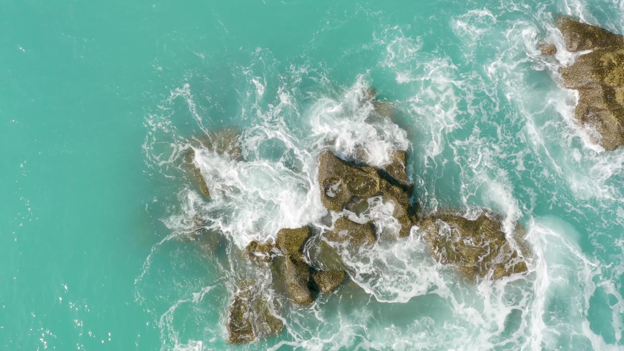 From above, white waves curl around rugged brown rocks in clear turquoise water at Dreams Beach, Baja California Sur, Mexico, with no visible people.