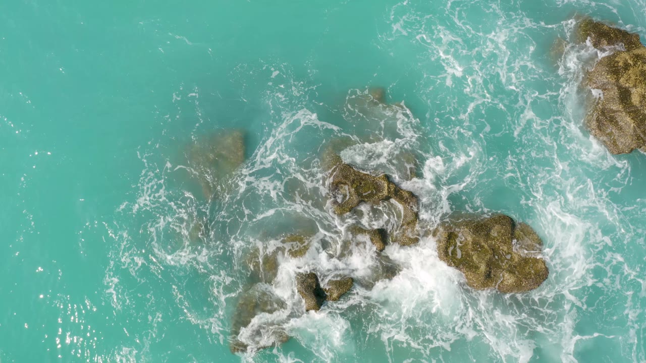 Aerial frame from Dreams Beach in Baja California Sur, Mexico, where foam wraps around the central rocks and a larger rock sits near the upper right, no visible people.