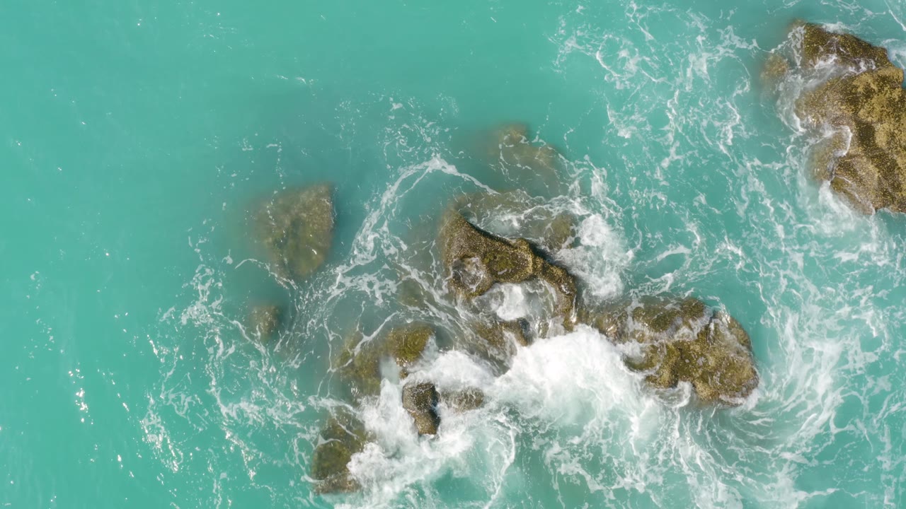 Overhead view at Dreams Beach in Baja California Sur, Mexico, with white wave wash spreading across the right side of the rock cluster and open turquoise water to the left, no visible people.
