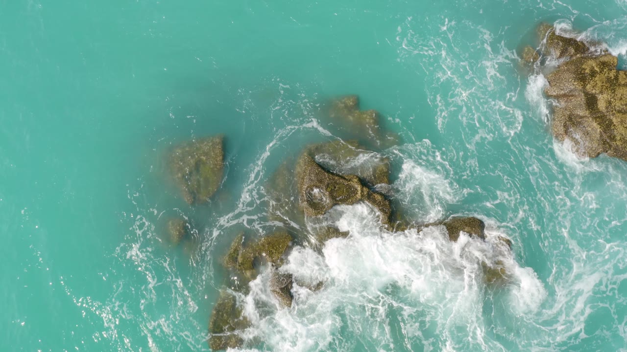 Aerial view at Dreams Beach in Baja California Sur, Mexico, showing the rock cluster lower in frame as foam pushes across the right edge and clear turquoise water opens at left, no visible people.