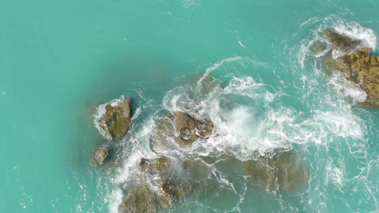 Aerial frame over Dreams Beach with white foam spreading through the center rocks and clear turquoise water around the edges, no visible people.