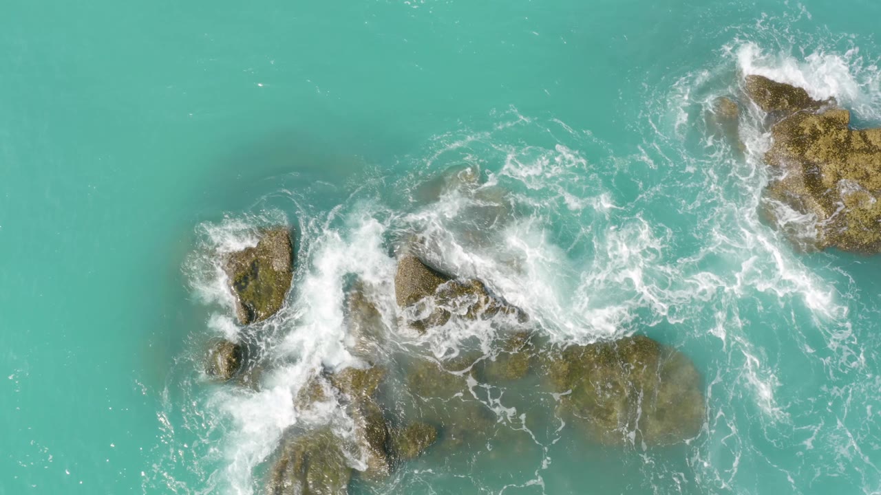 Whitewater crashes across the central rocks at Dreams Beach while larger brown stones sit at the upper right and blue-green water fills the background, no visible people.