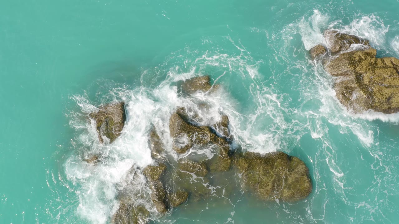 Overhead view of white surf wrapping around larger brown rocks near the lower frame at Dreams Beach, with turquoise water surrounding the formation and no visible people.