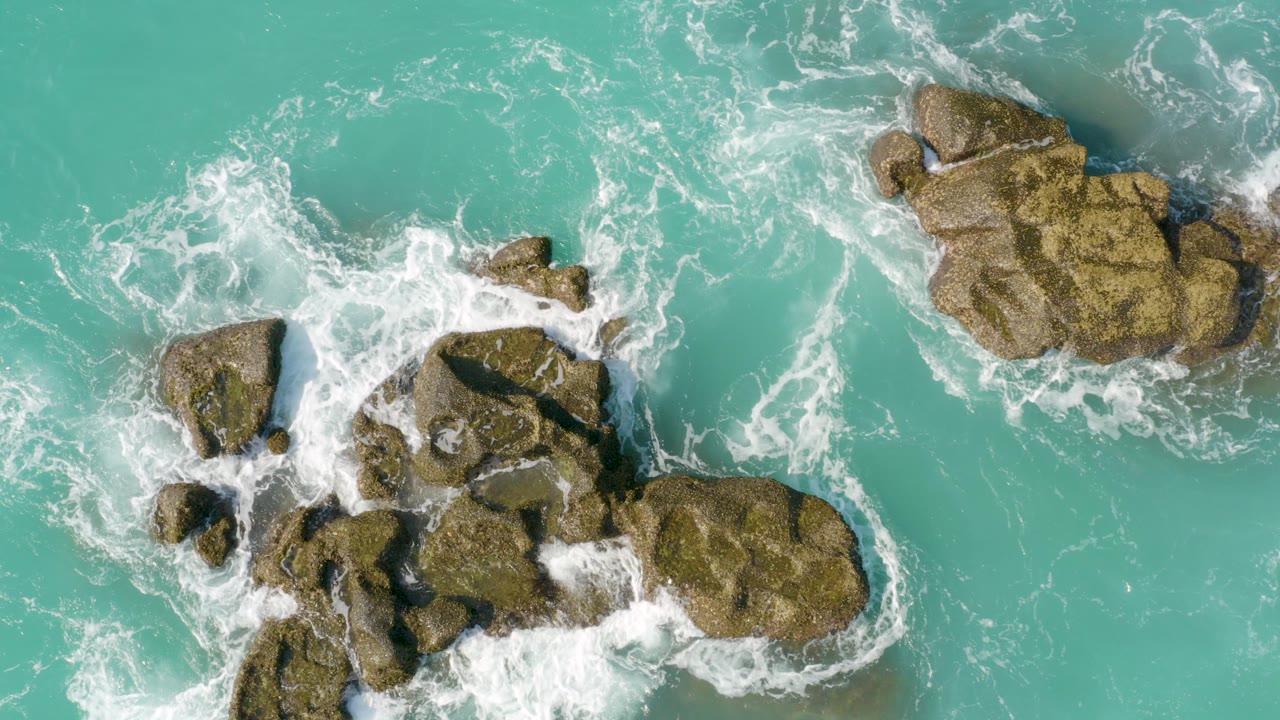 Overhead view at Dreams Beach, Baja California Sur, with turquoise water and white foam surrounding several tan rock clusters, no visible people.