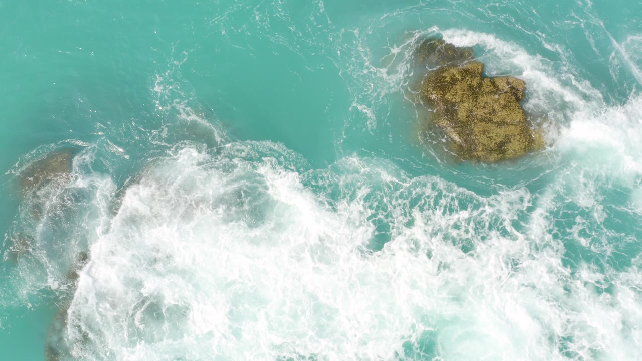 Turquoise surf breaking around rocks from above.