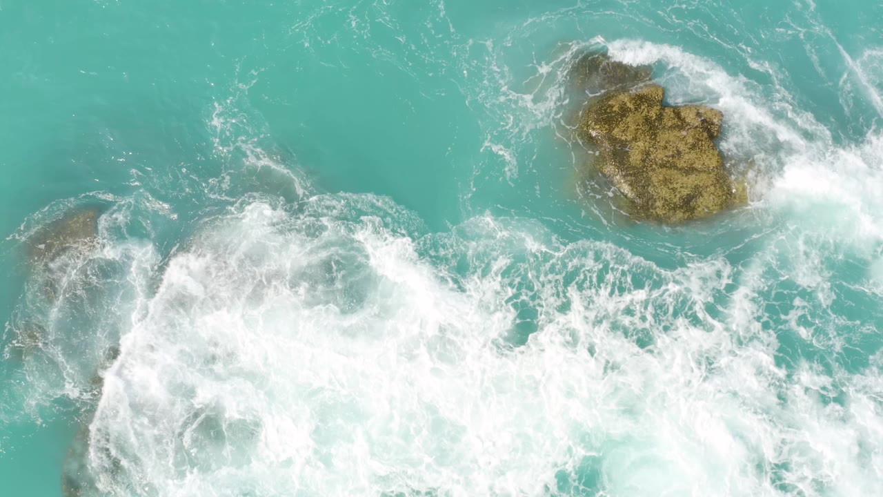 Aerial frame of blue-green water as a broader sheet of whitewater crosses the rocks at Dreams Beach in Baja California Sur, Mexico, with no visible people.