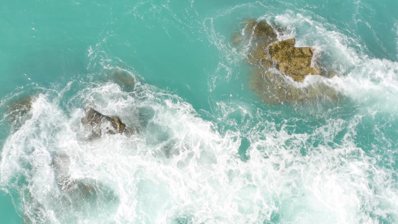 Top-down view of foamy surf breaking over scattered brown rocks in clear turquoise water at Dreams Beach in Baja California Sur, Mexico, with no visible people.