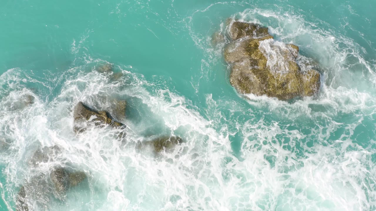 Overhead frame with several rocks emerging through white surf and pale blue water at Dreams Beach in Baja California Sur, Mexico, with no visible people.