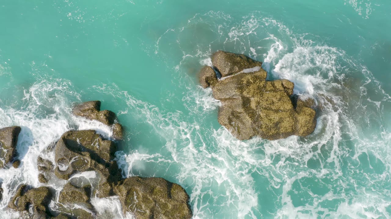 At 0:01, white foam spreads between a lower-left rock cluster and a larger upper rock in clear turquoise water at Dreams Beach, with no visible people.