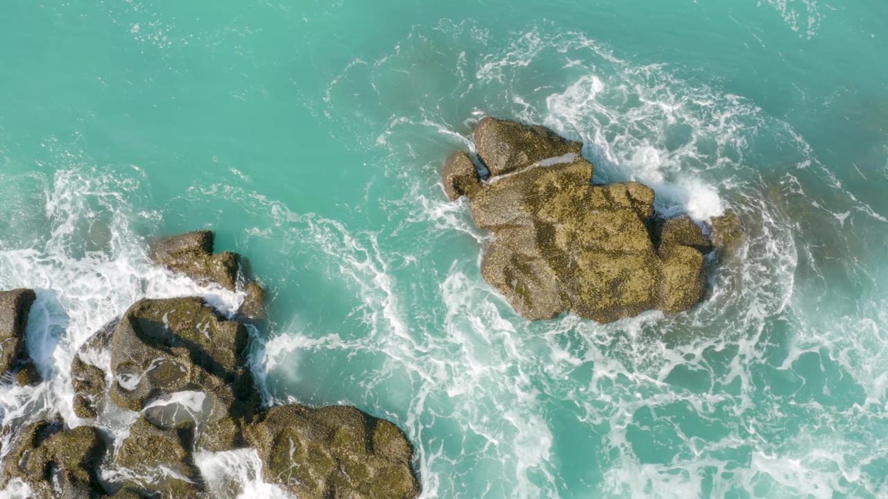 At 0:02, the central rock outcrop is ringed by breaking white water while smaller rocks sit lower in the frame at Dreams Beach, Baja California Sur, with no visible people.