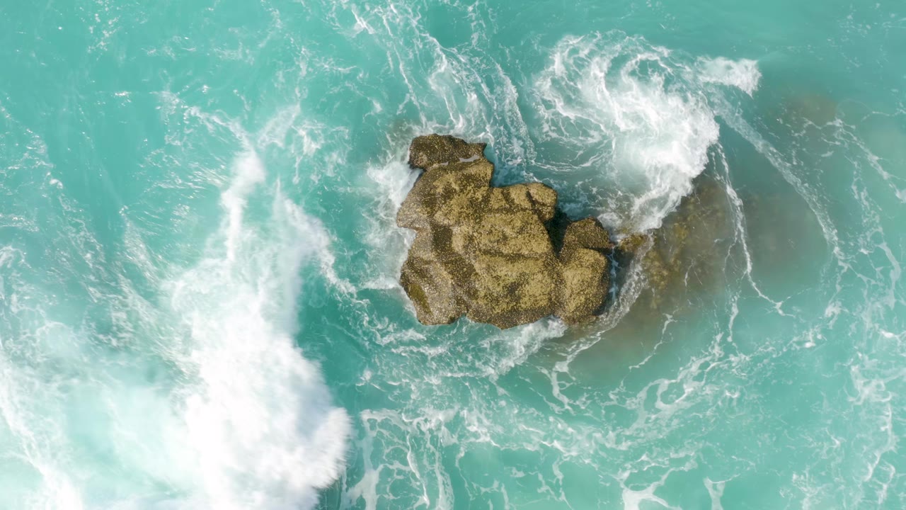 A top-down frame of white wave foam pressing in from the lower left toward a central tan rock in turquoise water at Dreams Beach, with no visible people.