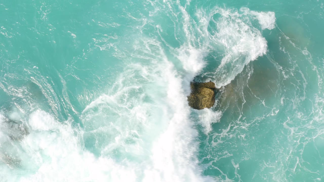 Whitewater spreads across the upper and left side of the frame while a smaller tan rock remains exposed in turquoise water at Dreams Beach, with no visible people.