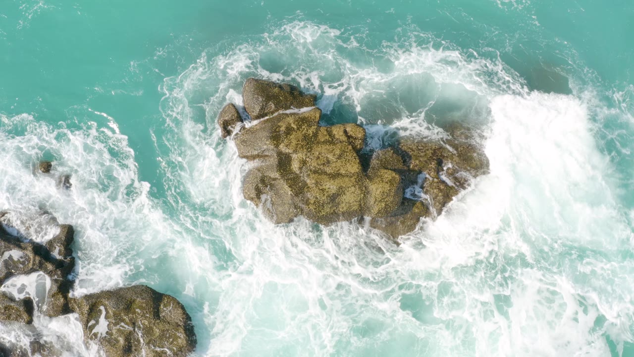 A stronger wave surge breaks against the upper right side of the brown rocks at Dreams Beach, Baja California Sur, with no visible people.