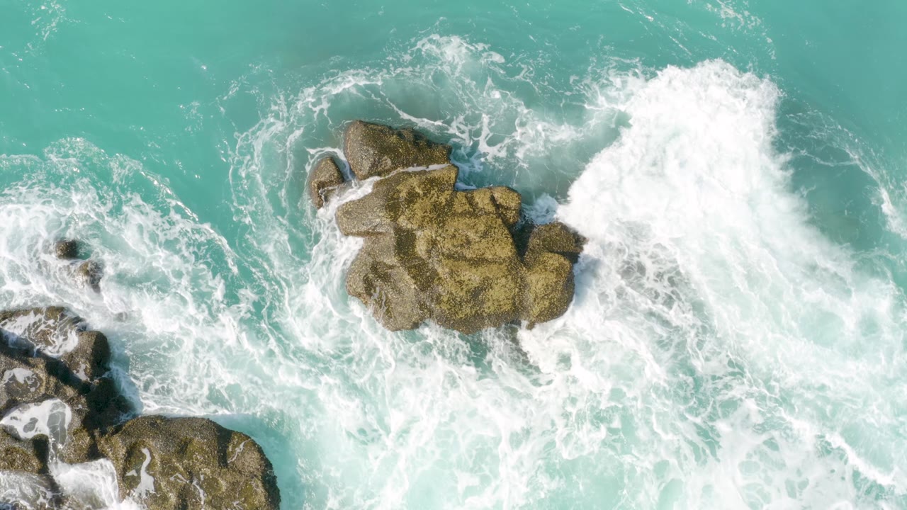 The overhead view shows the rock formation shifted lower in frame while foamy surf spreads across turquoise water at Dreams Beach, with no visible people.