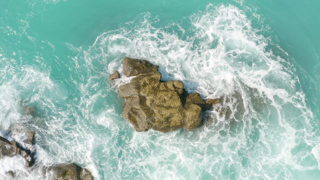 Overhead view at Dreams Beach in Baja California Sur, Mexico, with turquoise water and white foam surrounding tan rocks, no visible people.