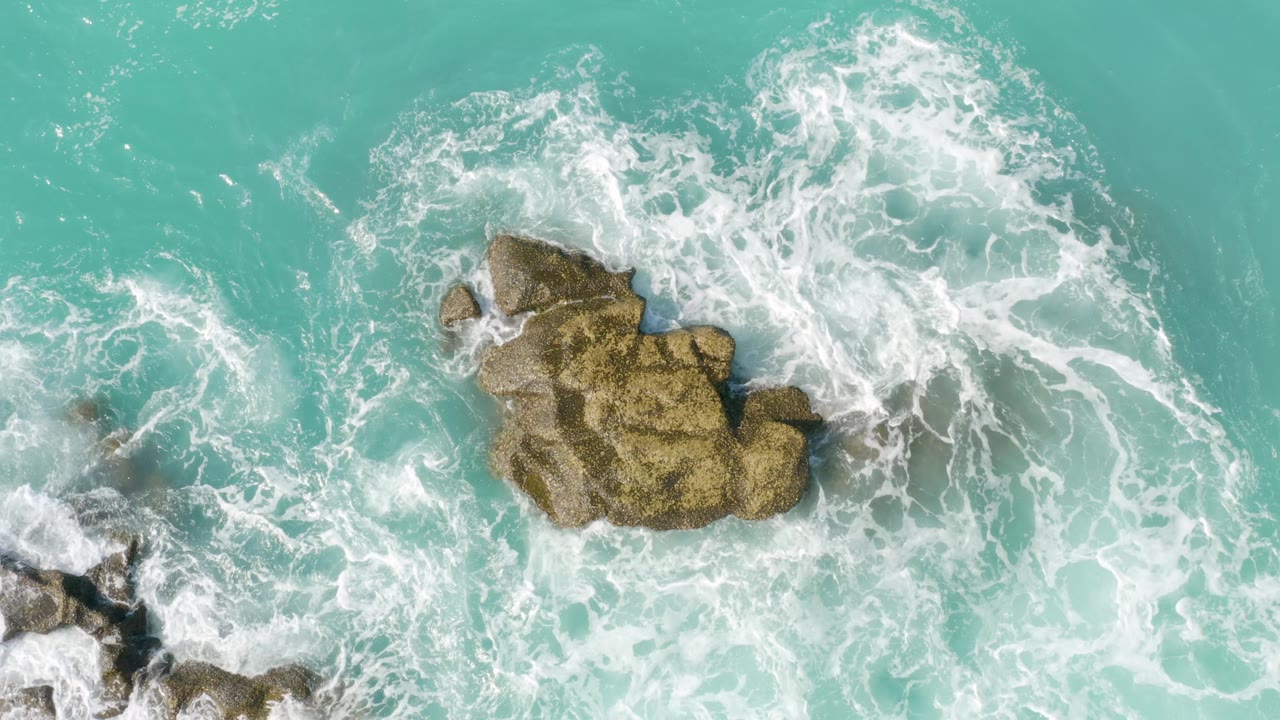 Overhead Dreams Beach view with a foamy wave breaking to the right of tan rocks in clear turquoise water, no visible people.