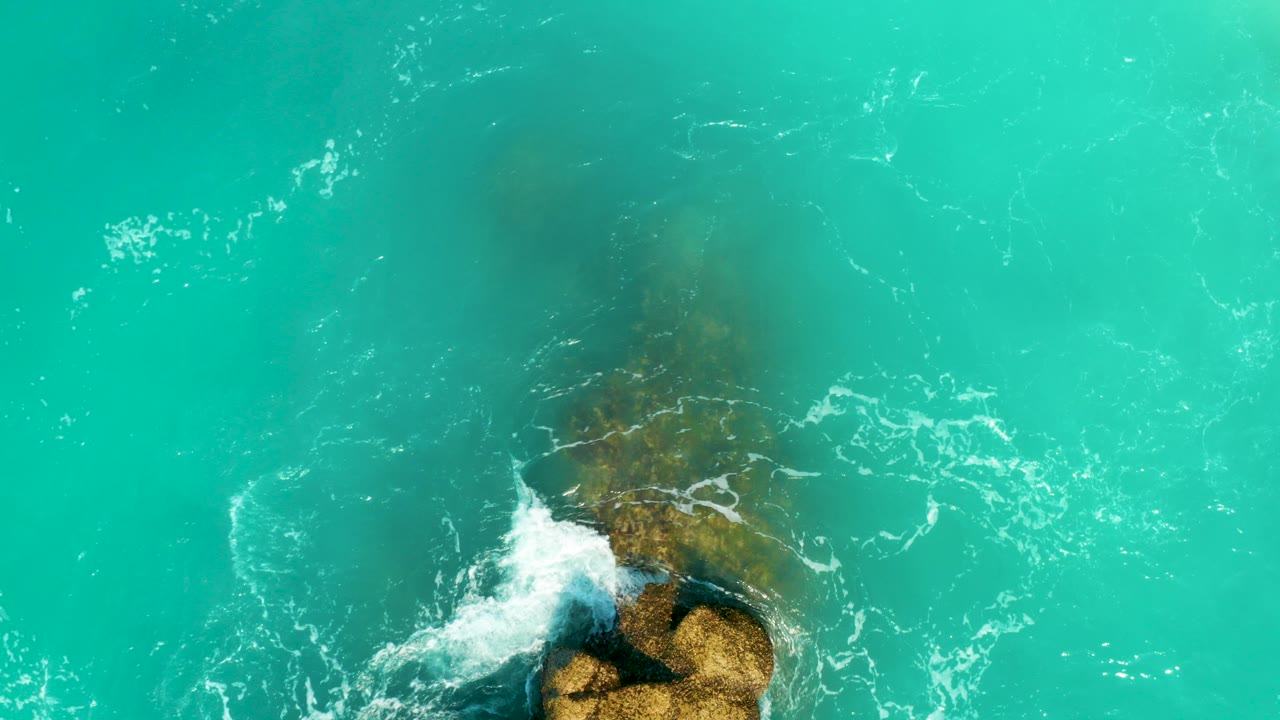 Overhead view at Dreams Beach in Baja California Sur, Mexico, showing turquoise water over a submerged brown rock as a small patch of white foam reaches it, with no visible people.