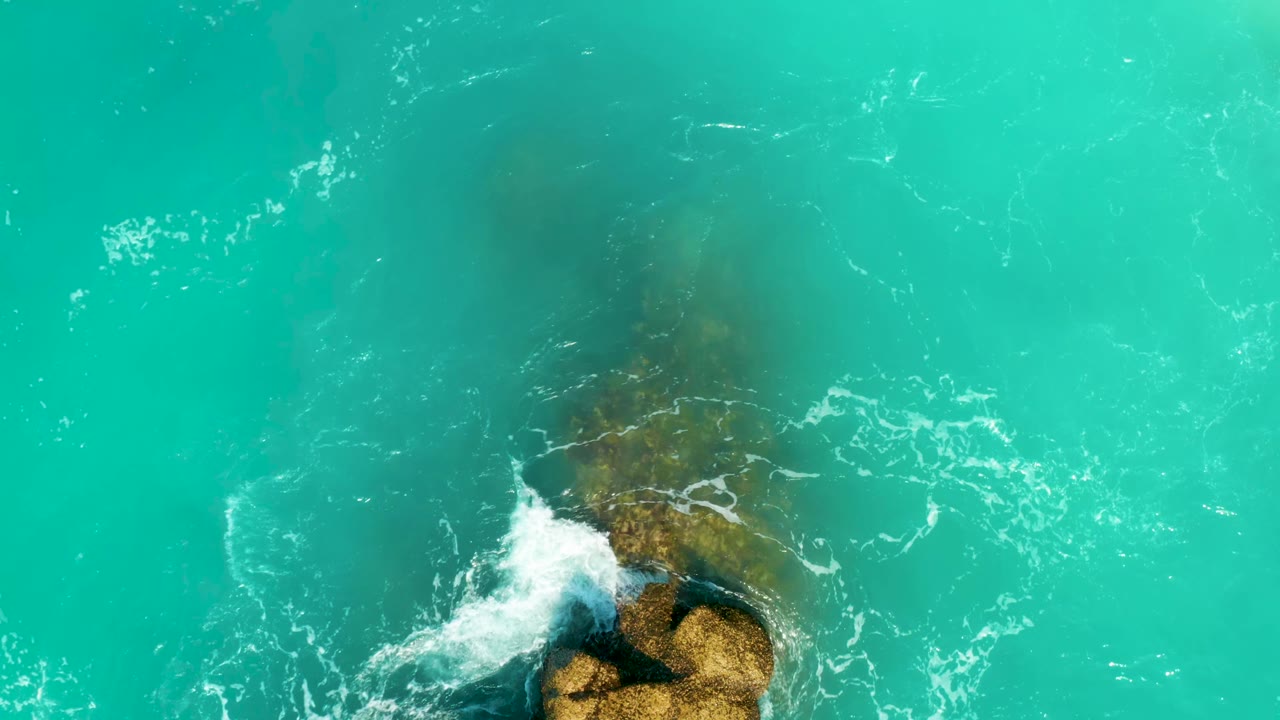 Overhead Dreams Beach frame with white surf starting to spread across the lower part of a submerged rock in turquoise water, with no visible people.