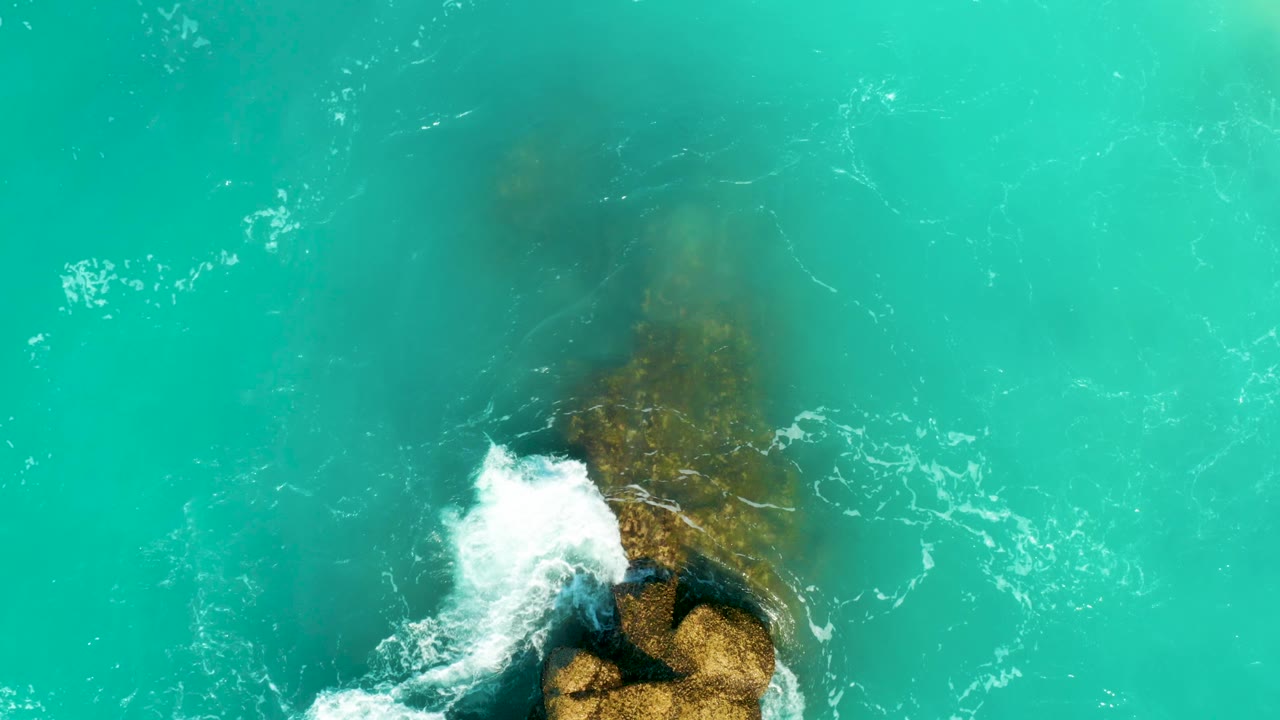 Aerial view of turquoise water at Dreams Beach as white wave foam climbs over the visible brown rock shape below the surface, with no visible people.