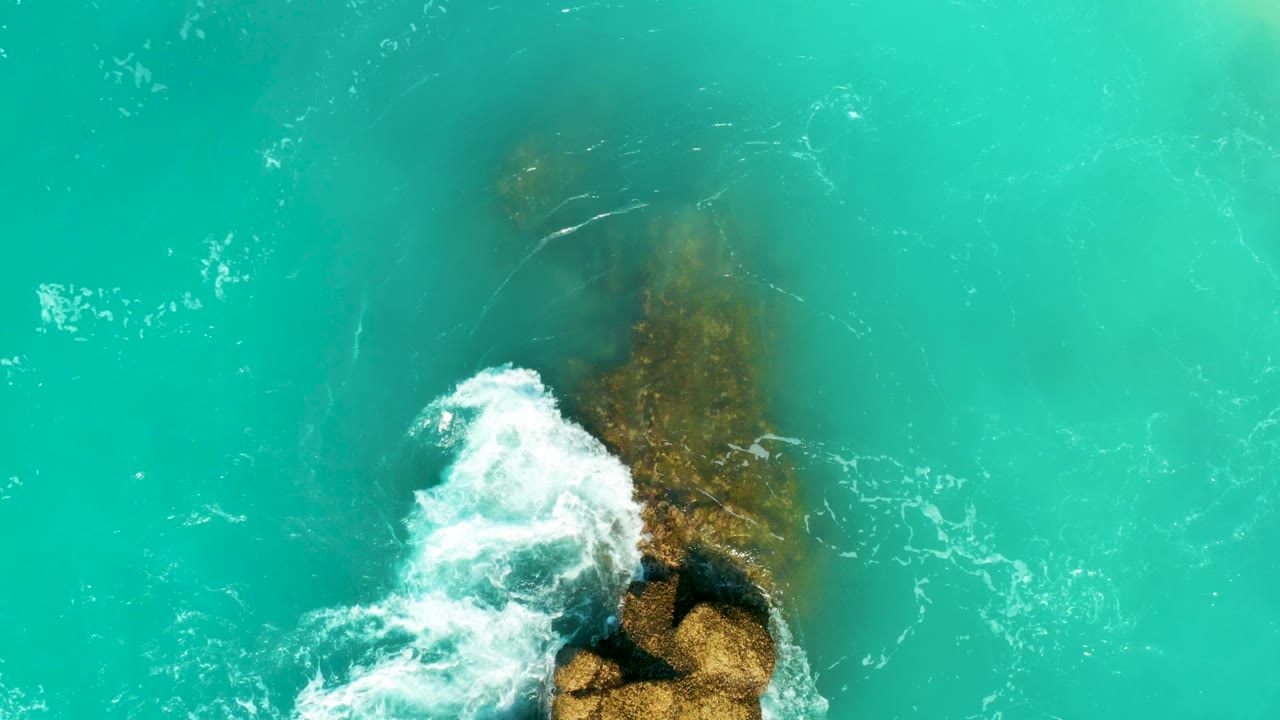Top-down frame from Dreams Beach with the submerged rock running vertically through turquoise water while white foam wraps around its lower edge, with no visible people.