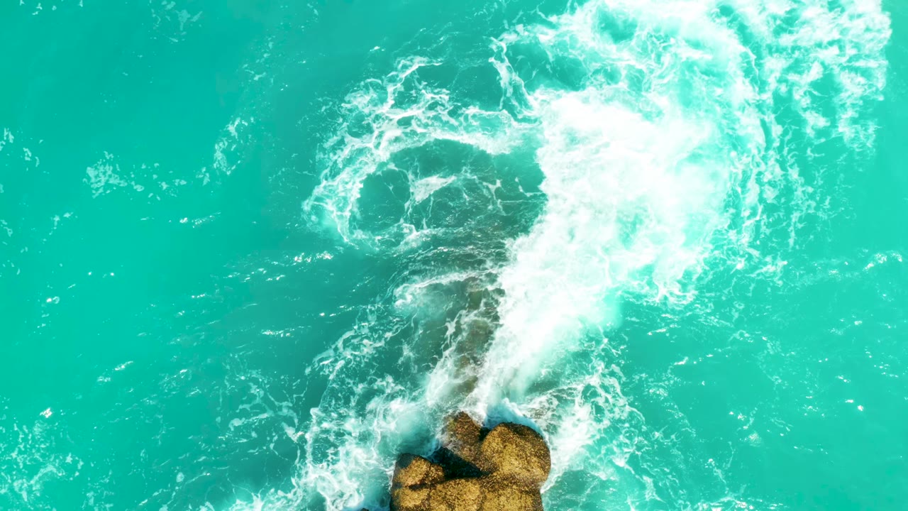 At 0:01, a thicker band of white foam curls diagonally through turquoise water while tan rocks remain near the bottom edge, with no visible people.