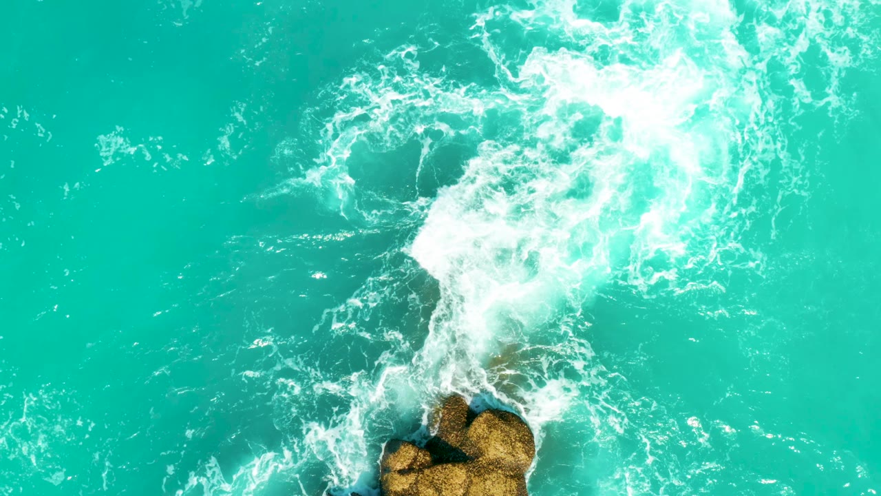 At 0:02, whitewater breaks from the upper right toward tan rocks at the bottom, with clear blue-green water filling the rest of the frame and no visible people.