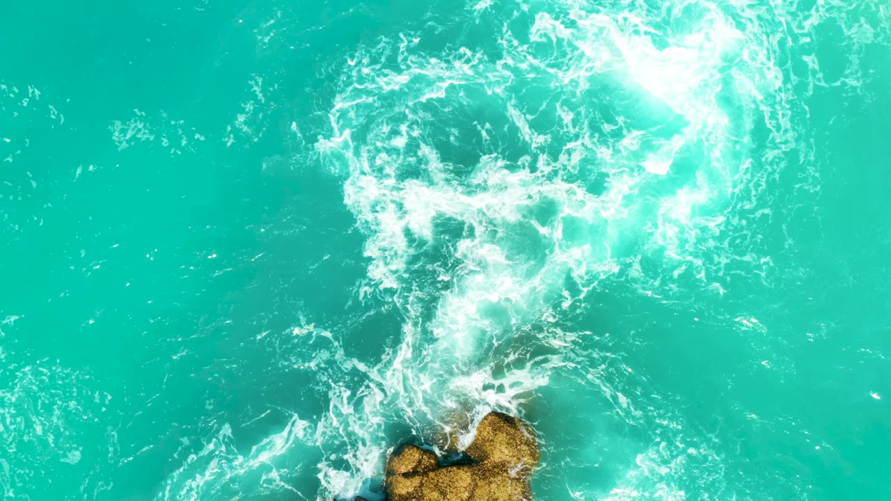 At 0:03, a long white foam trail runs upward from the lower rocks through turquoise water at Dreams Beach, with no visible people, boats, or buildings.