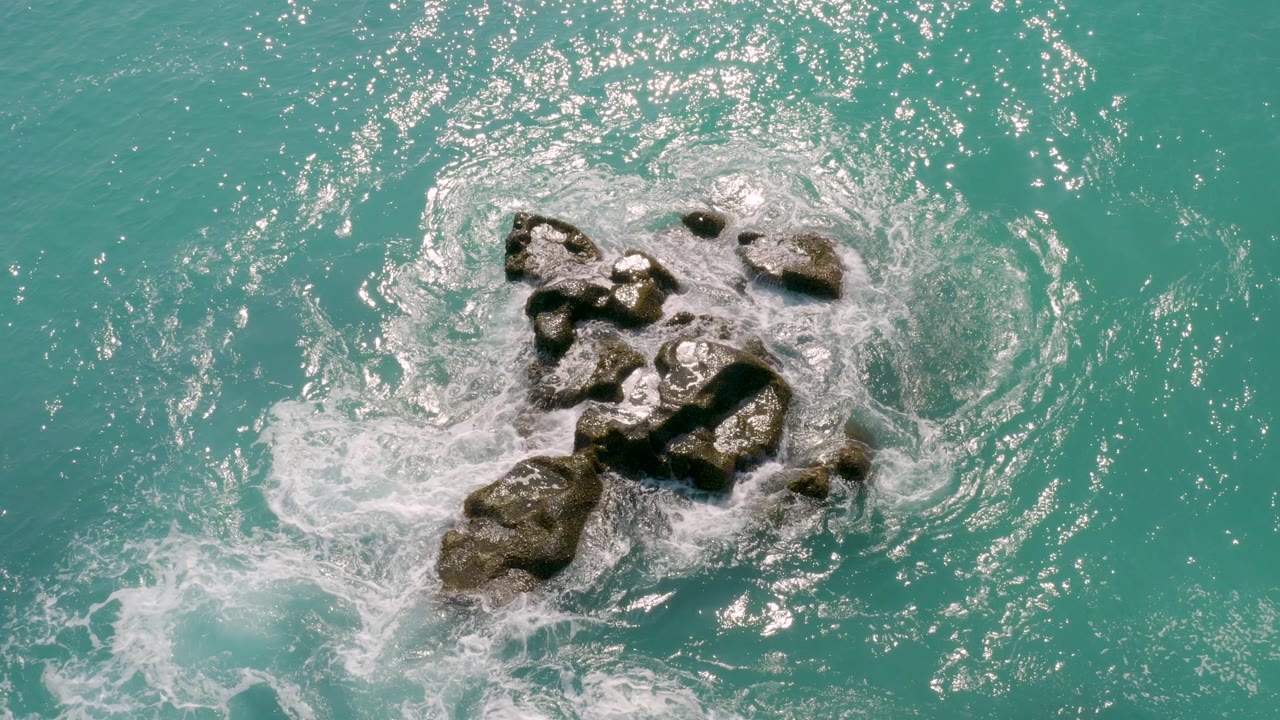 Overhead view at Dreams Beach in Baja California Sur showing turquoise water and white foam wrapping around a cluster of dark rocks, with no visible people.